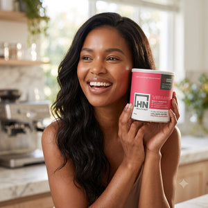 A smiling woman with long dark wavy hair holds a tub of HN Vibrance Collagen Powder in a bright kitchen setting.

